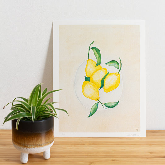 Image is a photo of a Limited edition art print sitting on bamboo wood surface with white background. The print is a reproduction from a handmade watercolour painting representing lemons with leaves on in a white bowl from above.