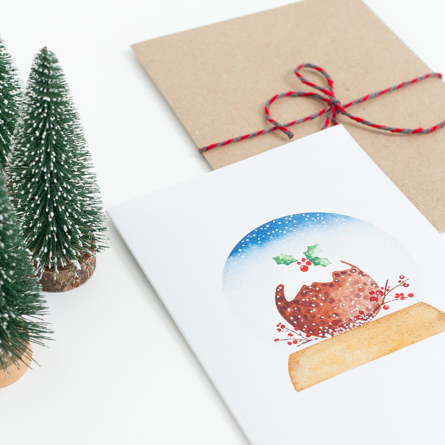 A festive food Snow Globe card designed from handmade watercolour painting. The illustration is a round Christmas pudding topped with icing and holly.