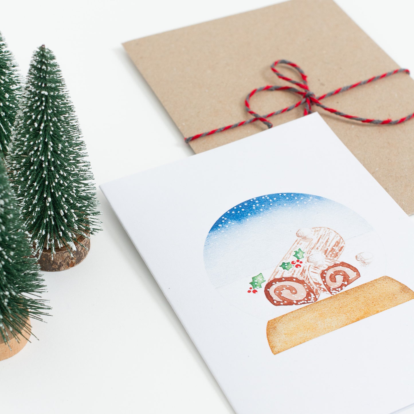 A festive food Snow Globe card designed from handmade watercolour painting. The illustration is a yule log (also known as bûche de Noël in France) topped with meringue mushrooms, cocoa powder and holly branches.