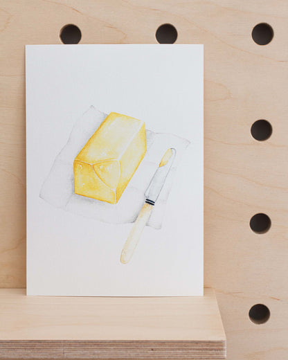 The art print is an illustration showing a block of butter with a piece taken out with a butter knife on the side. Created from handmade watercolour painting.