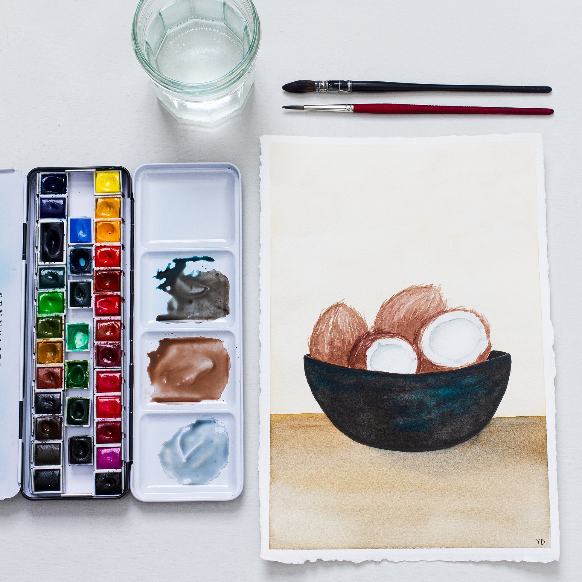 This photo shows an original painting of the coconut bowl with a palette of watercolour paints, two paint brushes and a small jar of water.