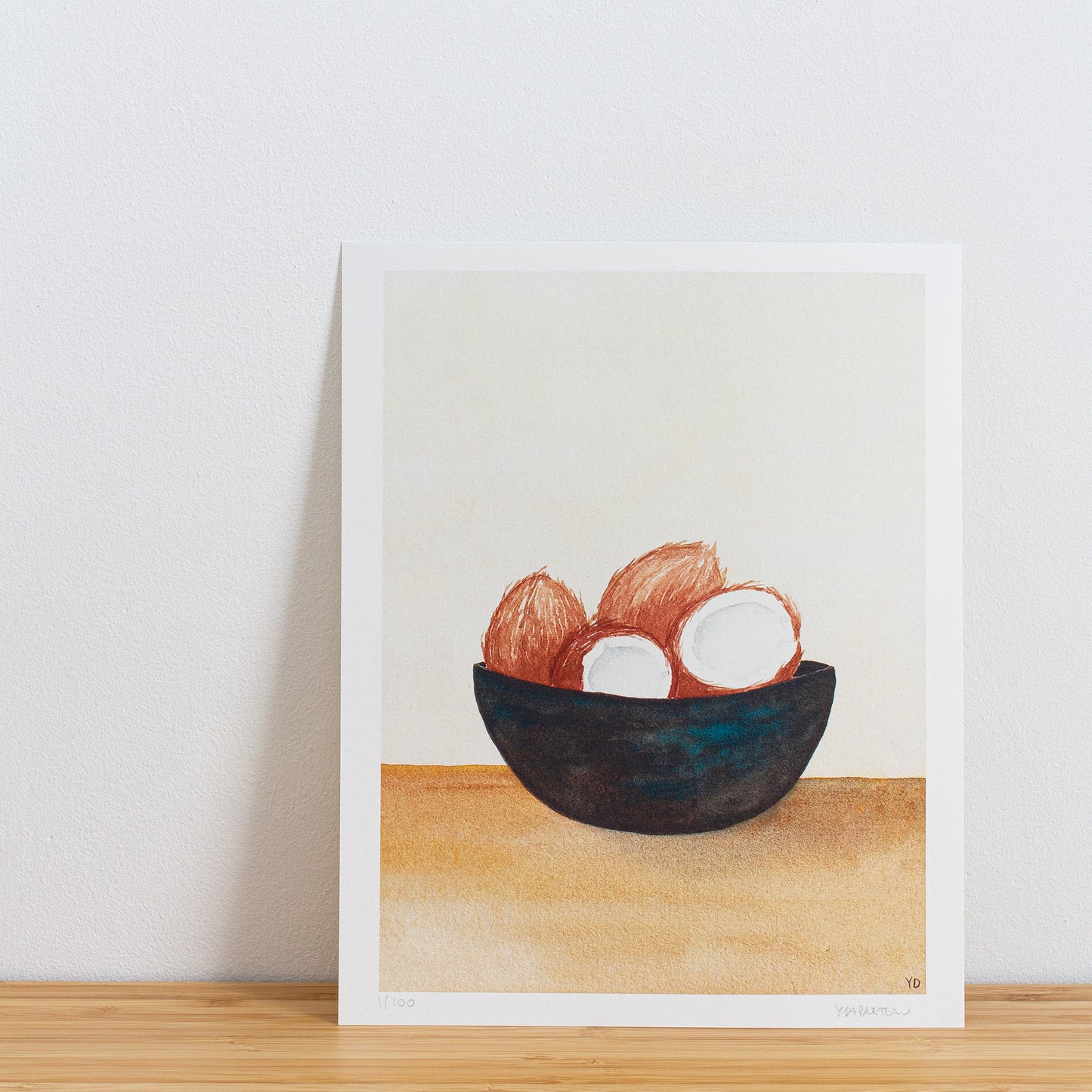 Image is a photo of a Limited edition art print sitting on bamboo wood surface with white background. The print is a reproduction from a handmade watercolour painting representing 4 coconuts in a dark bowl with turquoise streaks of paint throughout.