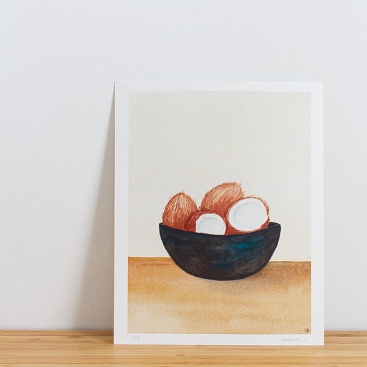 Image is a photo of a Limited edition art print sitting on bamboo wood surface with white background. The print is a reproduction from a handmade watercolour painting representing 4 coconuts in a dark bowl with turquoise streaks of paint throughout.