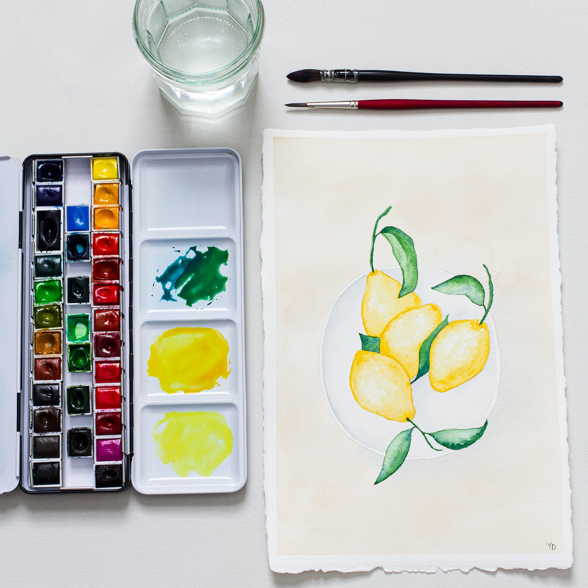 - This photo shows an original painting of the lemon bowl with a palette of watercolour paints, two paint brushes and a small jar of water.