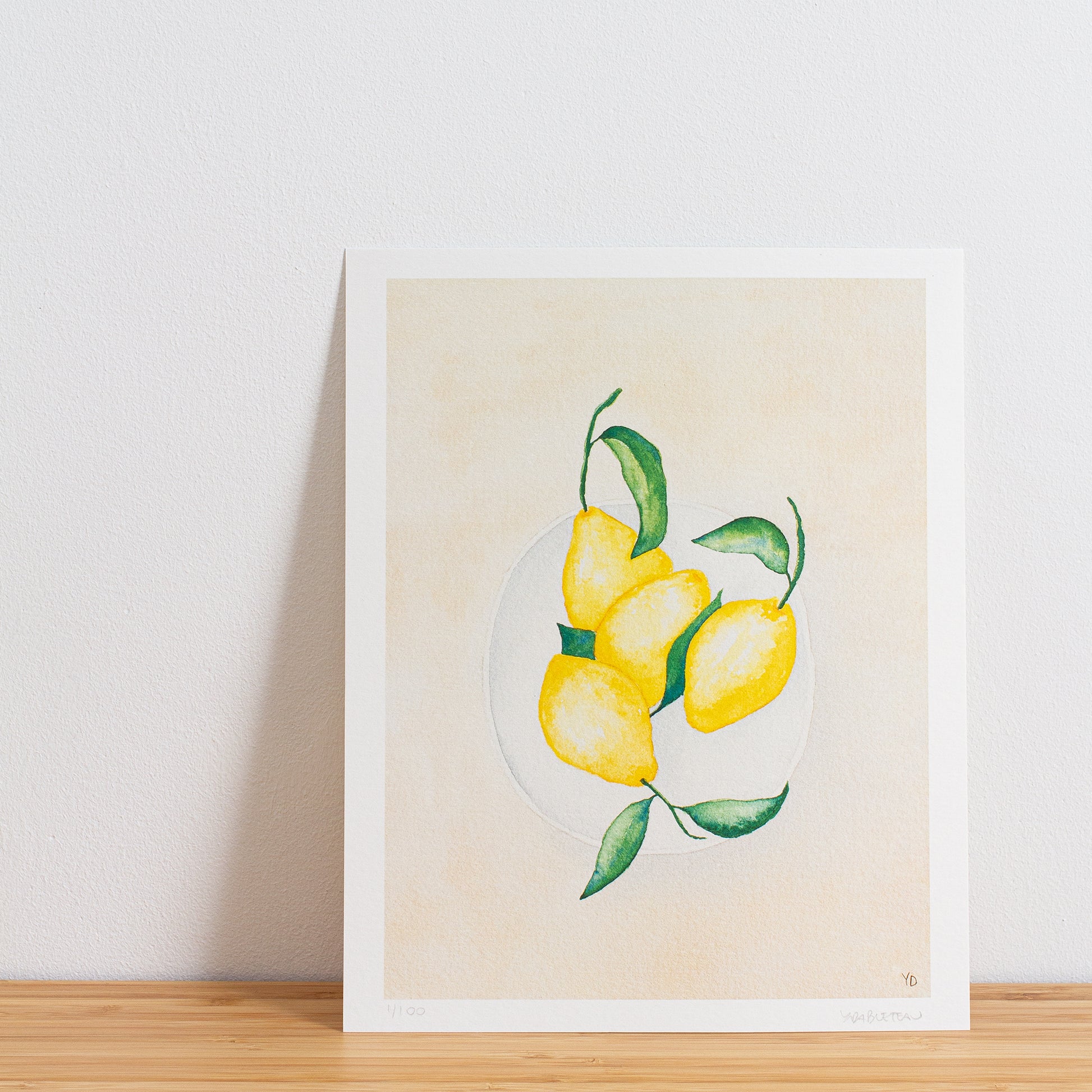 Image is a photo of a Limited edition art print sitting on bamboo wood surface with white background. The print is a reproduction from a handmade watercolour painting representing lemons with leaves on in a white bowl from above.