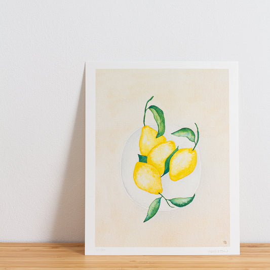 Image is a photo of a Limited edition art print sitting on bamboo wood surface with white background. The print is a reproduction from a handmade watercolour painting representing lemons with leaves on in a white bowl from above.