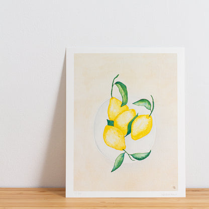 Image is a photo of a Limited edition art print sitting on bamboo wood surface with white background. The print is a reproduction from a handmade watercolour painting representing lemons with leaves on in a white bowl from above.