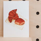 The art print is an illustration showing two croissants and a pain au chocolat on a piece of paper. Created from handmade watercolour painting.