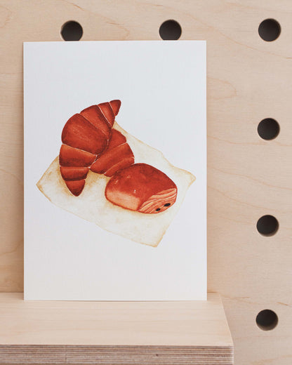 The art print is an illustration showing two croissants and a pain au chocolat on a piece of paper. Created from handmade watercolour painting.