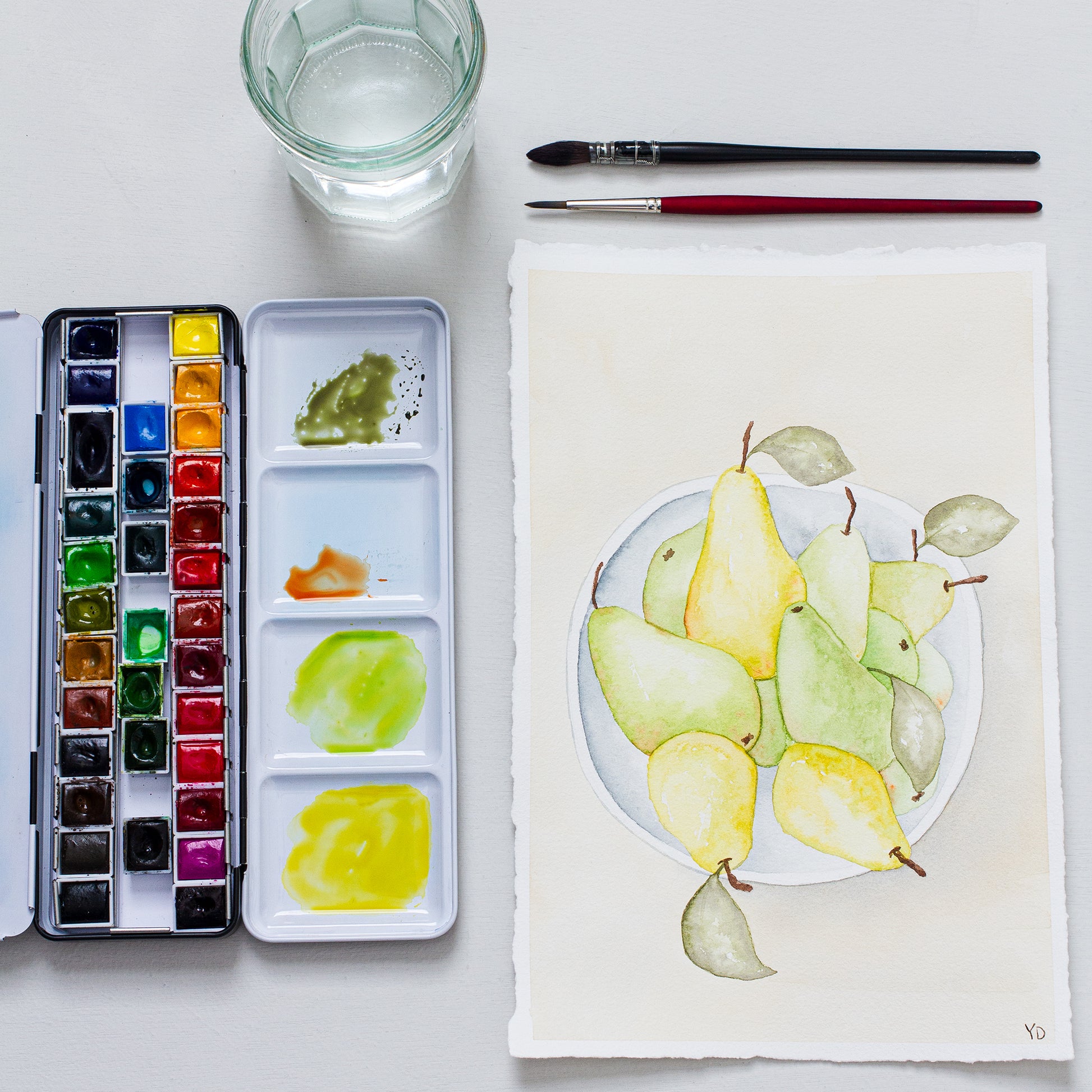 This photo shows an original painting of the pear bowl with a palette of watercolour paints, two paint brushes and a small jar of water.