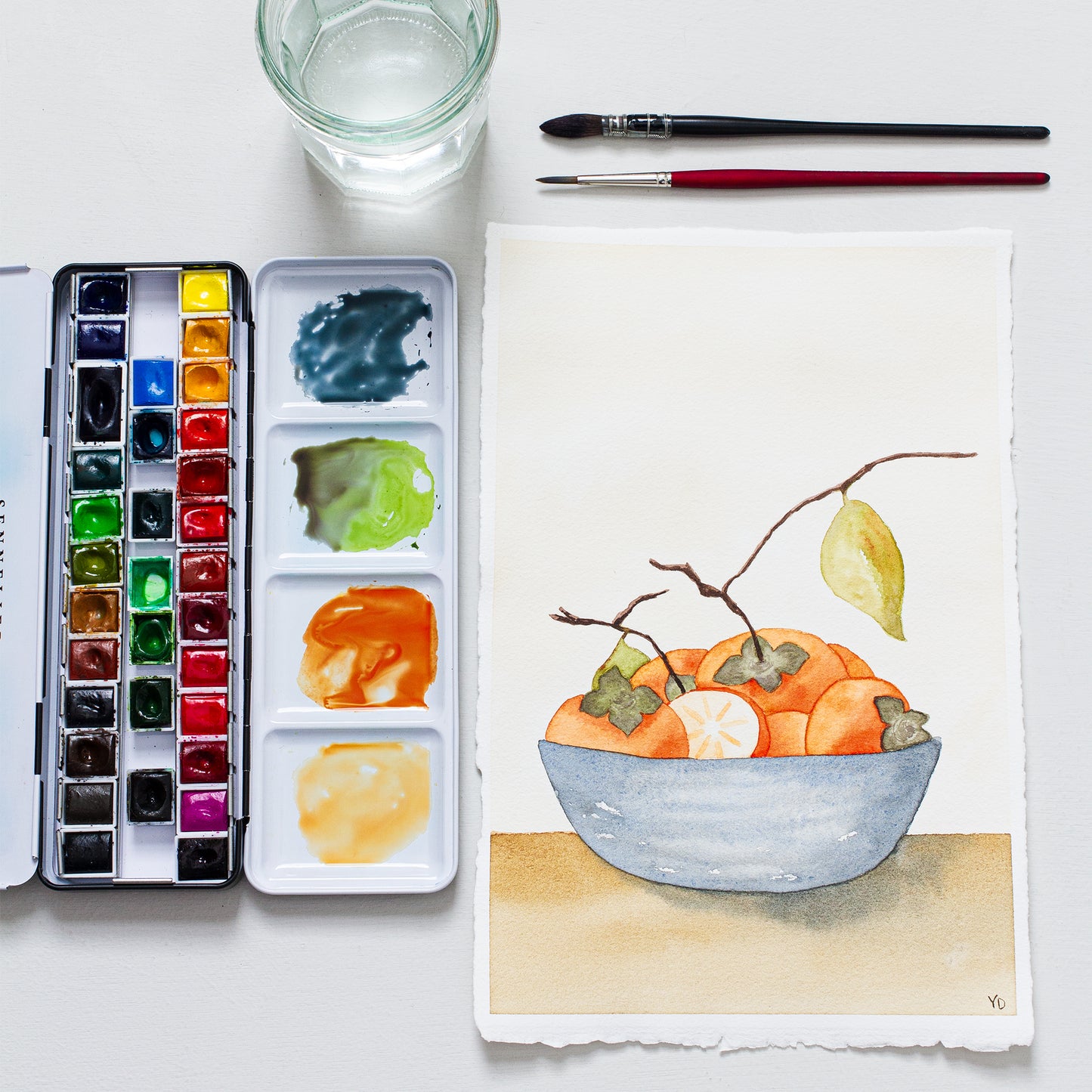 This photo shows an original painting of the persimmon bowl with a palette of watercolour paints, two paint brushes and a small jar of water.