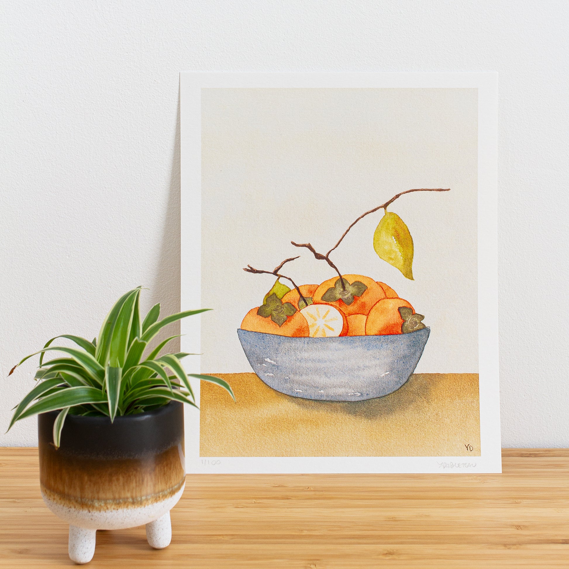 Image is a photo of a Limited edition art print sitting on bamboo wood surface with white background. The print is a reproduction from a handmade watercolour painting representing persimmon fruits with branches and leaves in a blue grey bowl.