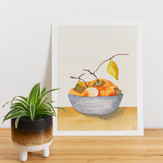Image is a photo of a Limited edition art print sitting on bamboo wood surface with white background. The print is a reproduction from a handmade watercolour painting representing persimmon fruits with branches and leaves in a blue grey bowl.