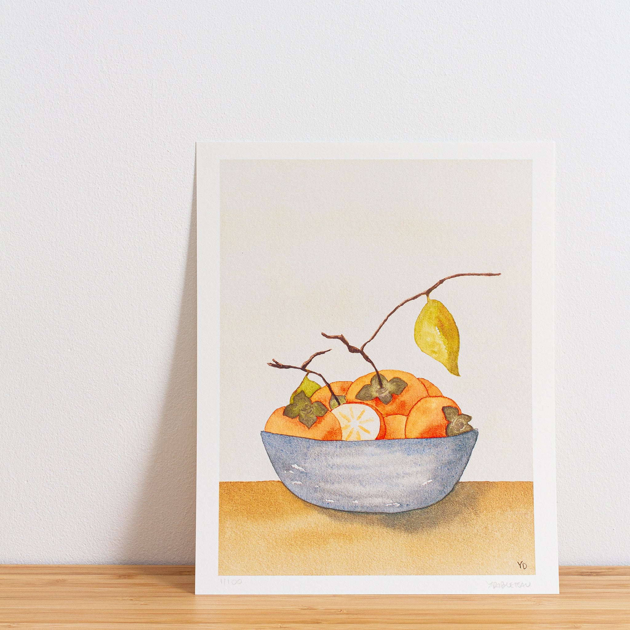 Image is a photo of a Limited edition art print sitting on bamboo wood surface with white background. The print is a reproduction from a handmade watercolour painting representing persimmon fruits with branches and leaves in a blue grey bowl.
