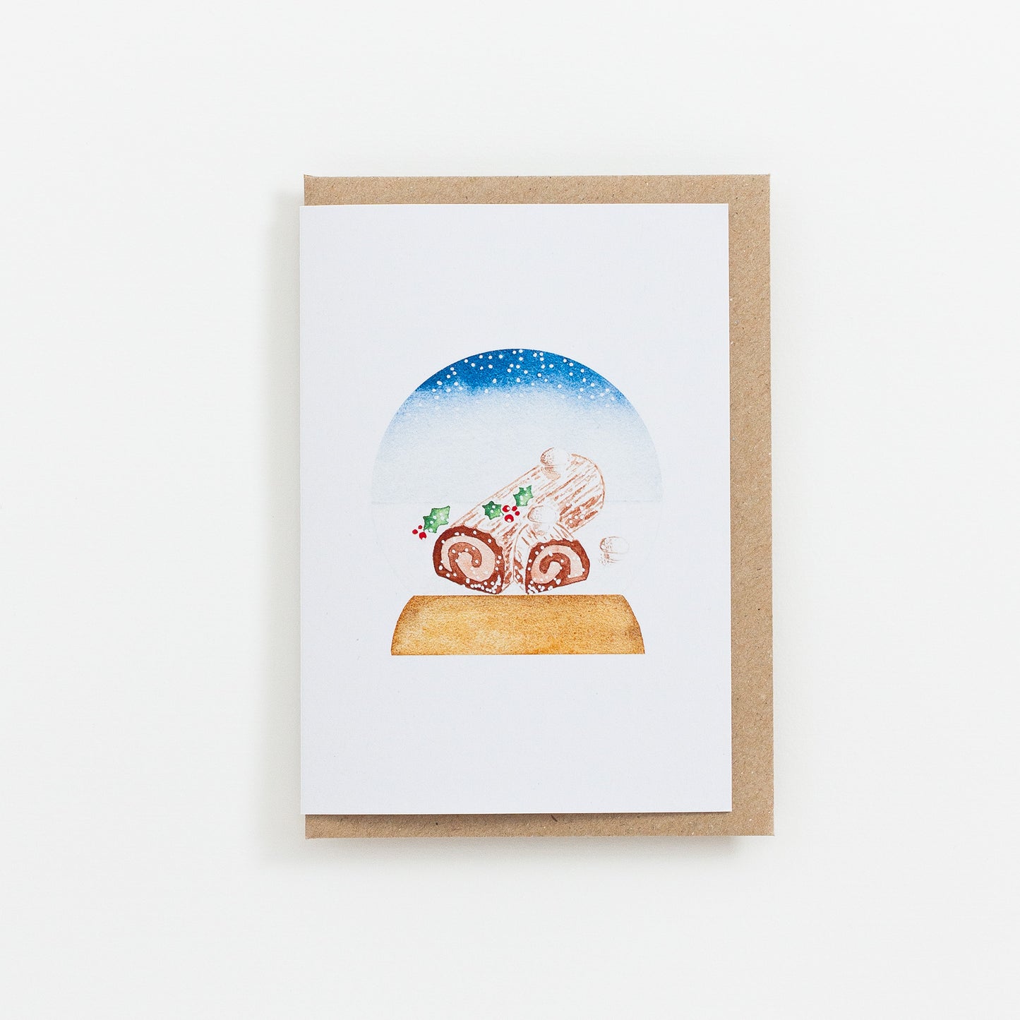 A festive food Snow Globe card designed from handmade watercolour painting. The illustration is a yule log (also known as bûche de Noël in France) topped with meringue mushrooms, cocoa powder and holly branches.