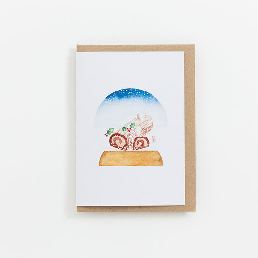 A festive food Snow Globe card designed from handmade watercolour painting. The illustration is a yule log (also known as bûche de Noël in France) topped with meringue mushrooms, cocoa powder and holly branches.